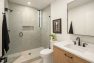 A photo of the interior of a modern new construction townhome. Shows the primary bathroom with focus on the fully tiled walk-in shower with frameless glass door, built-in shelf, and a frosted window for natural light.