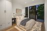 A photo of the interior of a modern new construction townhome. Focuses on the primary bedroom with a sliding glass door that leads to a large balcony. Includes furnishings such as a built-in wardrobe cabinet, two black wooden nightstands, a large bed with upholstered headboard, brass reading lamps, and a large jute rug.