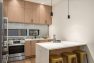 A photo of the interior of a modern new construction townhome. Focuses on the kitchen with a large white kitchen island with seating. Features woodgrain floors and cabinetry, stainless steel kitchen appliances, simple black hardware, and a glossy gray-blue subway tile backsplash.