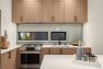 A photo of the interior of a modern new construction townhome. Focuses on the kitchen with two windows. Features woodgrain floors and cabinetry, stainless steel kitchen appliances, simple black hardware, and a glossy gray-blue subway tile backsplash.