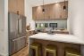 A photo of the interior of a modern new construction townhome. Focuses on the kitchen with a large white kitchen island with seating. Features woodgrain floors and cabinetry, stainless steel kitchen appliances, and simple black hardware and pendant lighting.