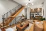 A photo of the interior of a modern new construction townhome. Focuses on the living room and dining space with a prominent, modern wood and metal staircase. Includes furnishings such as a camel leather sofa, a woodgrain rectangular coffee table, and a round dining table with four bent metal dining chairs in gold finish.