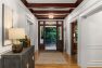 An image of the interior of a 1905 Neoclassical home. Focuses on the formal entry hall with a view of the open front door, leading out to the entry porch. Includes hardwood floors, detailed white millwork and traditional wainscoting, decorative leaded glass clerestory windows above the door, and a wooden beam ceiling.