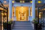 An image of the exterior of a 1905 Neoclassical home. Focuses on the approach to the formal, main entry door. Includes a wrought-iron decorative gate, large potted plants, and a view of the main-level entry porch as well as the second-level balcony above. Features architectural elements common to Neoclassical style such as fluted and ornamented columns.