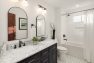 An image of the interior of a 1905 Neoclassical home. Focuses on a guest bathroom in a traditional style with a white and black color palette. Features a black, double-sink vanity cabinet with white and gray marble countertop, black and white faucets, and two black-framed mirrors. Also includes traditional styled overhead sconces, tiled floors, and a white, subway tiled shower-bath combo.