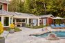 An image of a residential exterior, with an in-ground pool in the foreground. Focuses on accessory structures to a private home, including an open-air cabana with gray outdoor sofas and a wooden dining table.