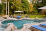 An image of a residential backyard surrounded by mature trees. Focuses on an in-ground swimming pool with waterfall and rock-like surrounding patio. Includes wicker outdoor lounge seating and white umbrellas.