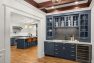 An image of the interior of a 1905 Neoclassical home. Focuses on the built-in wine bar and cabinetry with a partial view into the chef's kitchen.. Features detailed millwork, wood beam ceiling, hardwood floors, semi-flushmount glass light fixtures and globe pendant light fixtures.