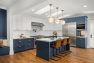 An image of the interior of a 1905 Neoclassical home. Focuses on the spacious chef's kitchen with a blue and white color palette. Features multiple skylights, hardwood floors, a large kitchen island with sink, a double oven and large gas range, globe pendant lights, and brown leather barstools.