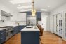 An image of the interior of a 1905 Neoclassical home. Focuses on the spacious chef's kitchen with a blue and white color palette. Features multiple skylights, hardwood floors, a large kitchen island with sink, a double oven and large gas range, globe pendant lights, and brown leather barstools.