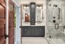 An image of the interior of a 1905 Neoclassical home. Focuses on the primary bathroom in a gray, white, bronze and gold color palette. Features a custom double-sink vanity with floor to ceiling cabinetry and two large, rectangular mirrors. Also includes a fully tiled, walk-in shower with built-in bench seating and glass door. Floors are tiled in a herringbone pattern. Tile are natural stone with rich gray veining.